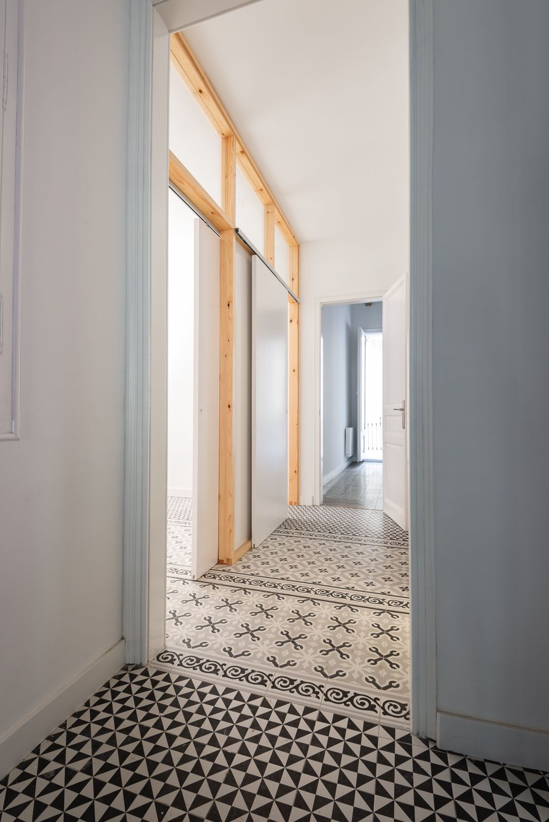 Hallway with patterned floor tiles and wooden frame elements leading to a bright room.