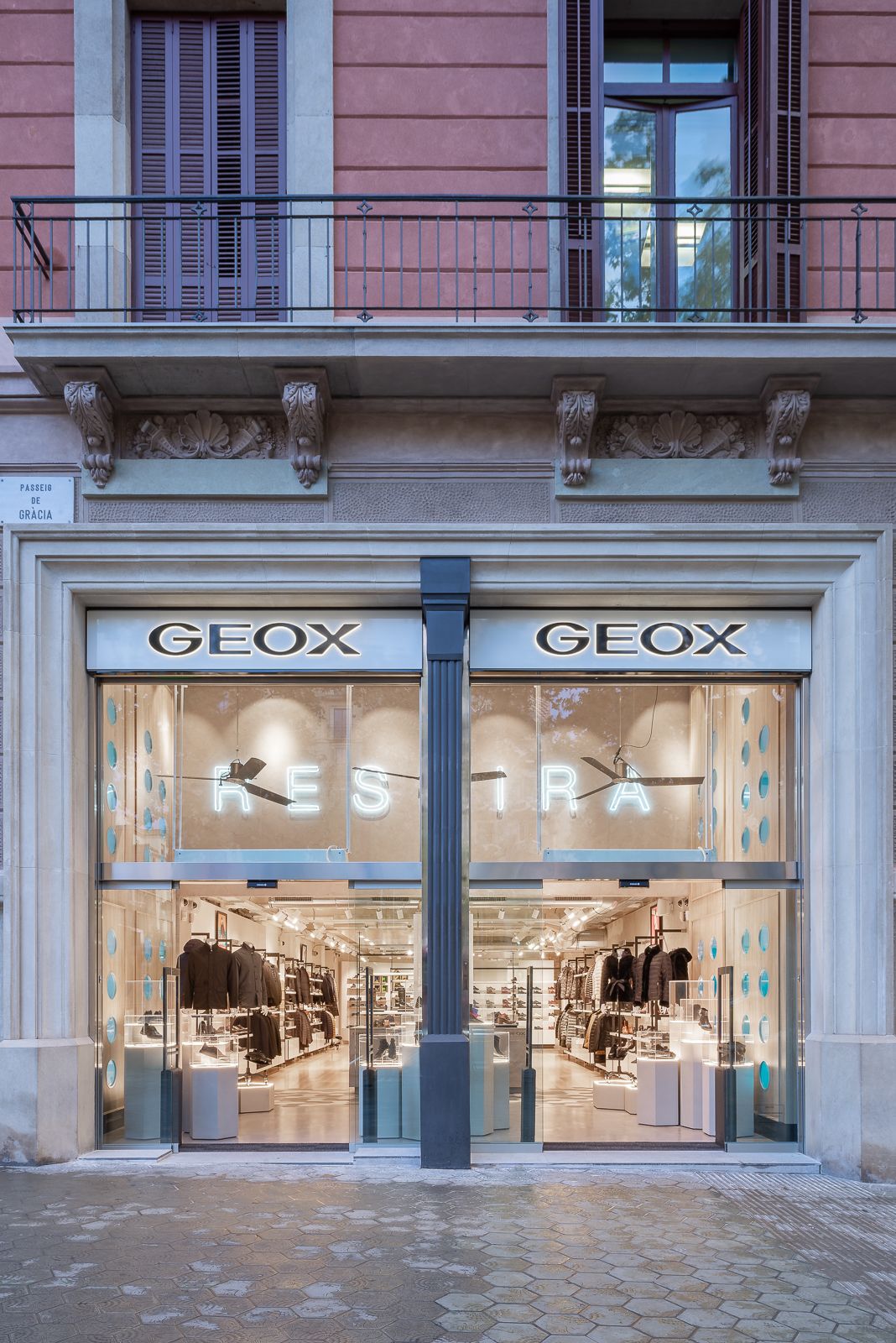 Vista frontal d'una botiga Geox al Passeig de Gràcia a Barcelona, amb grans finestres de vidre que mostren la mercaderia interior i un balcó a sobre.