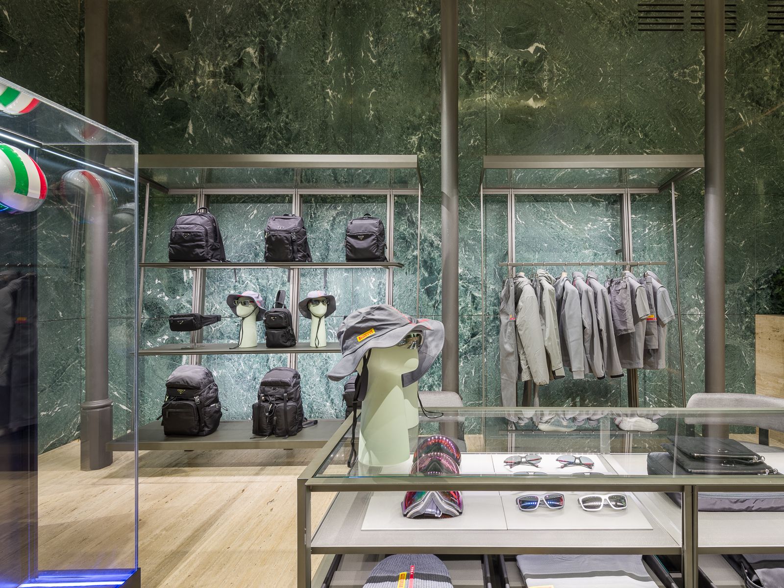 Luxury boutique with green marble walls, showcasing designer sneakers, black bags, and accessories on sleek metal shelving. A glass table displays eyewear and hats, while a mannequin in a performance outfit is encased in a glass display.