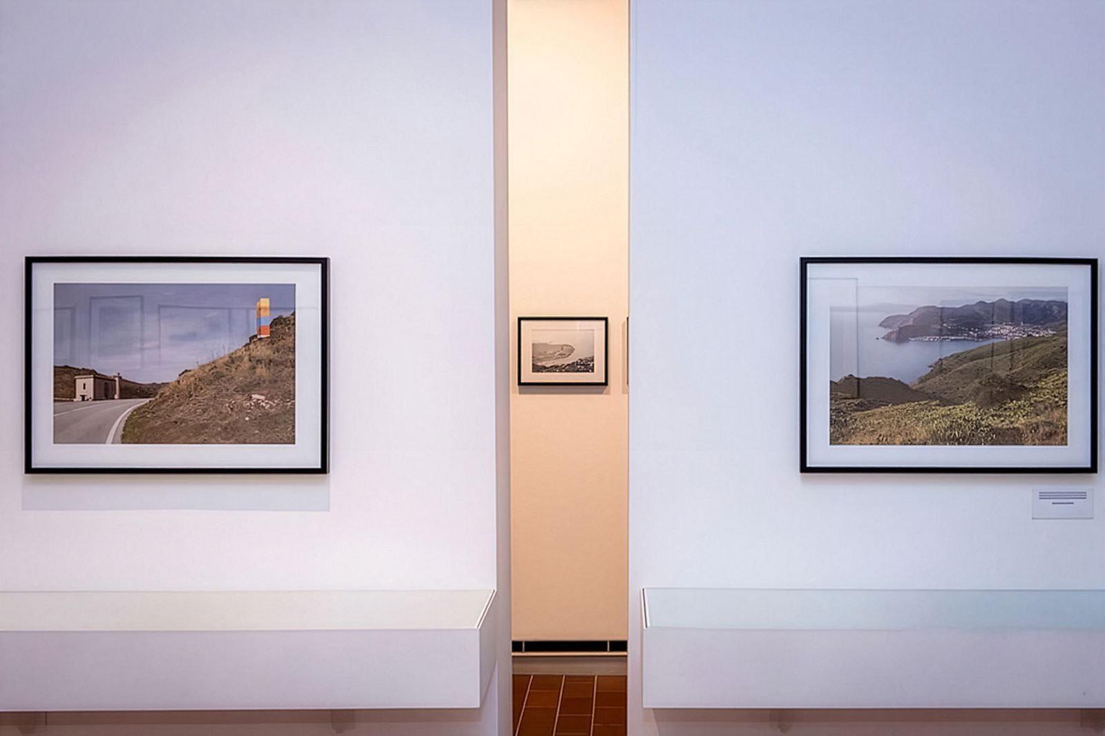 Dos fotografías de paisaje enmarcadas se muestran en una pared blanca con una pequeña fotografía en blanco y negro visible al fondo a través de una abertura.