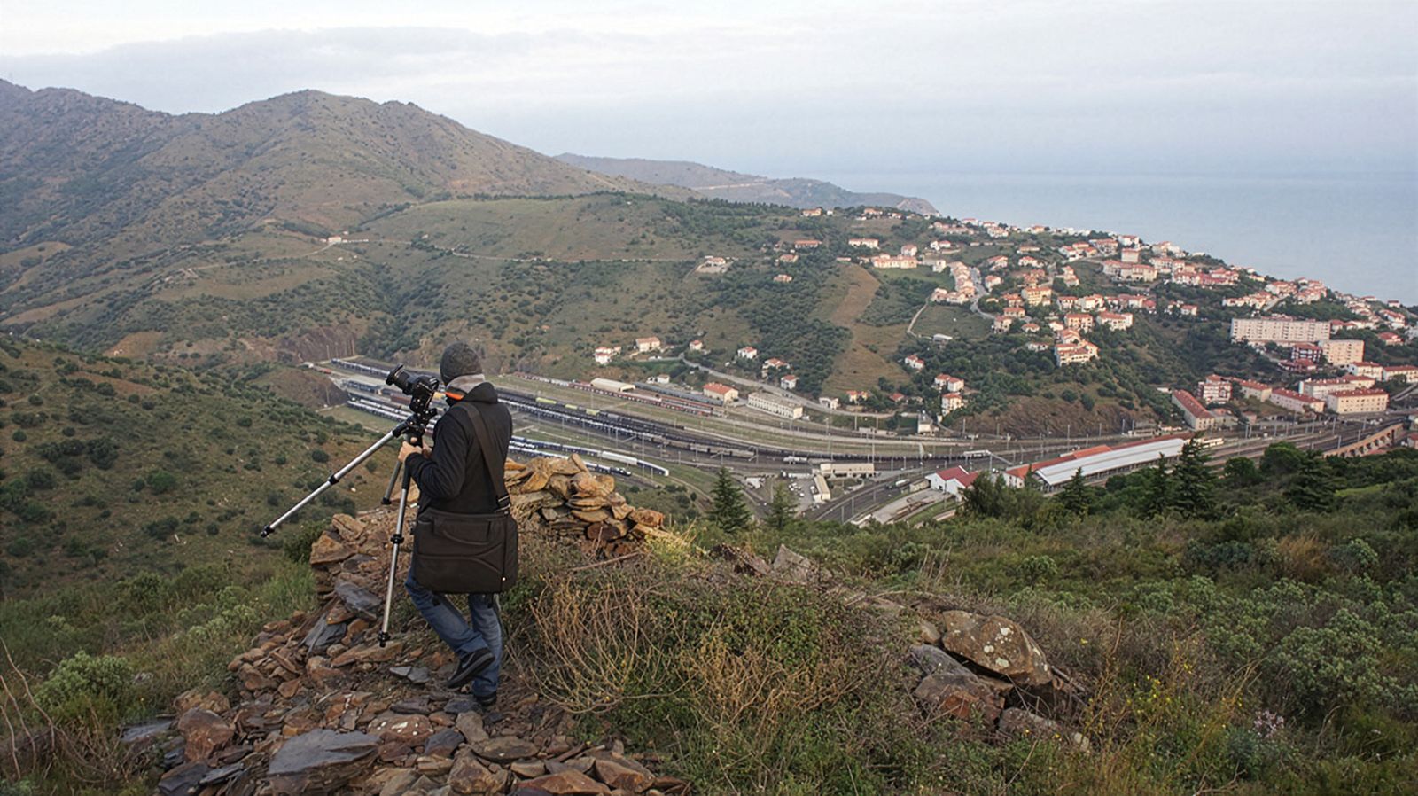 Un fotógrafo con un trípode a punto de capturar una vista panorámica de un pueblo costero con una gran estación de tren enclavada entre colinas.