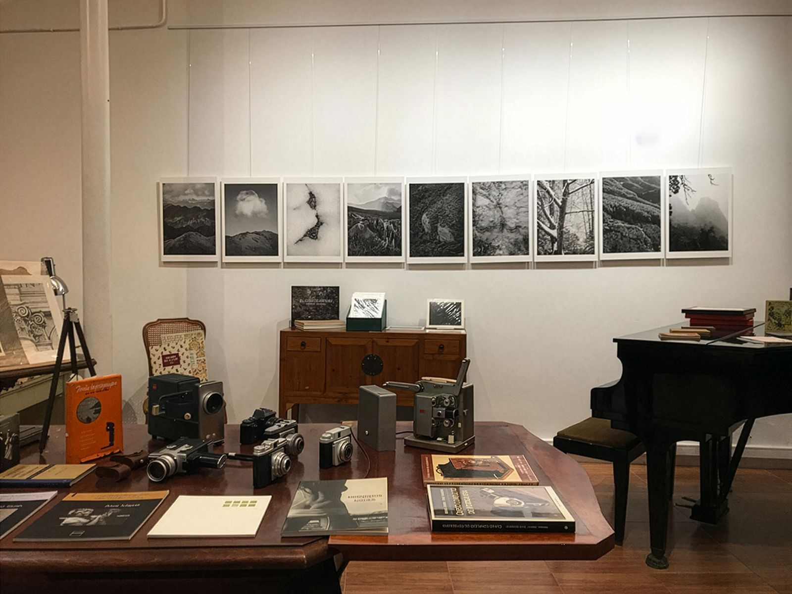 Un espai d'exposició amb fotografies en blanc i negre de muntanyes en una paret, una taula amb càmeres antigues i llibres de fotografia, un piano i diversos altres llibres i objectes exposats.