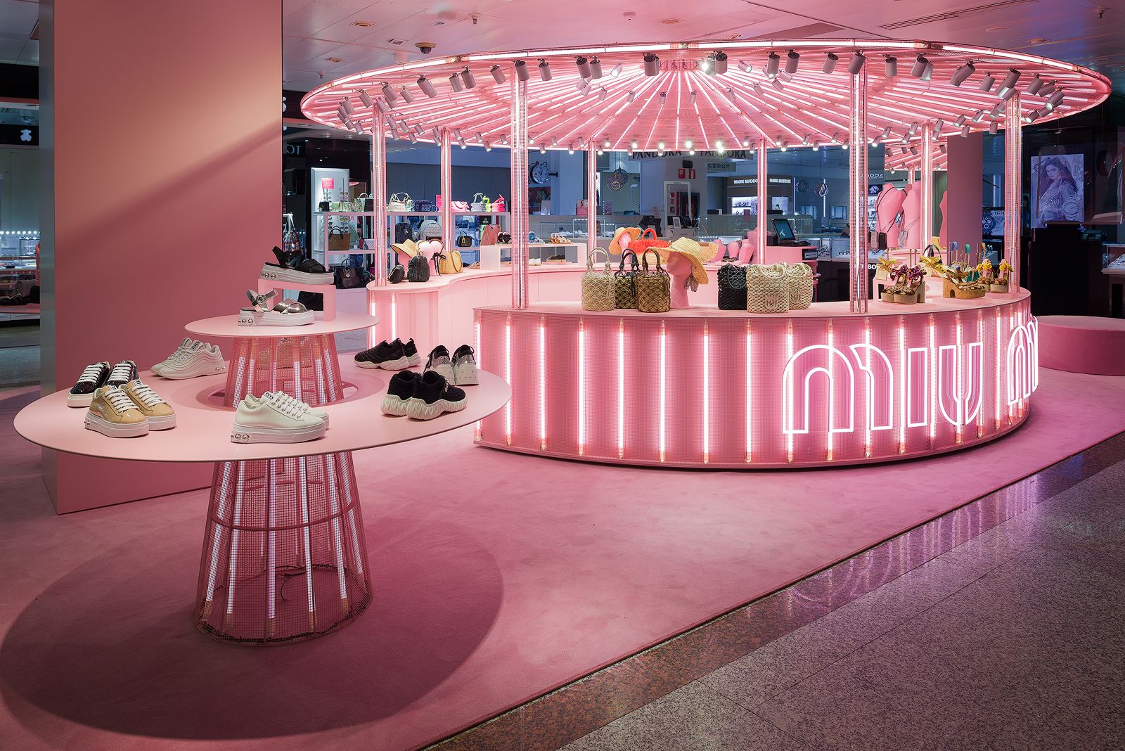 Pink-themed Miu Miu pop-up store display with illuminated round tables showcasing sneakers, handbags, and accessories.