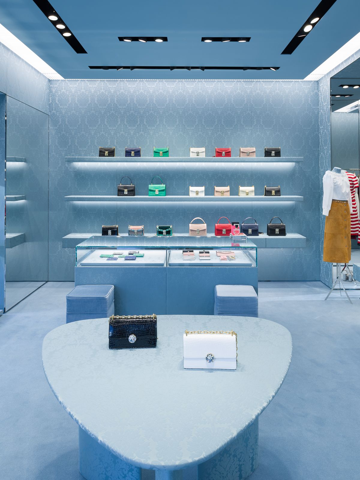 Miu Miu store interior with blue patterned walls, black lighting fixtures, and colorful handbags displayed on sleek shelves and glass counters.
