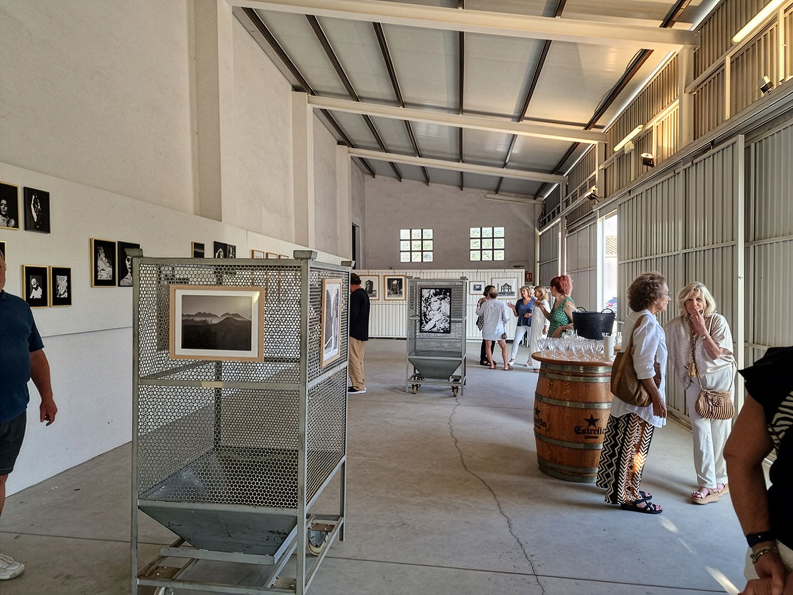 Interior de un espacio de exposición con techos altos y vigas expuestas. Las paredes muestran fotografías enmarcadas. Se ven soportes de exhibición metálicos y un barril de madera. Varios visitantes están de pie y conversando en pequeños grupos por todo el espacio, dando la impresión de una inauguración de galería.