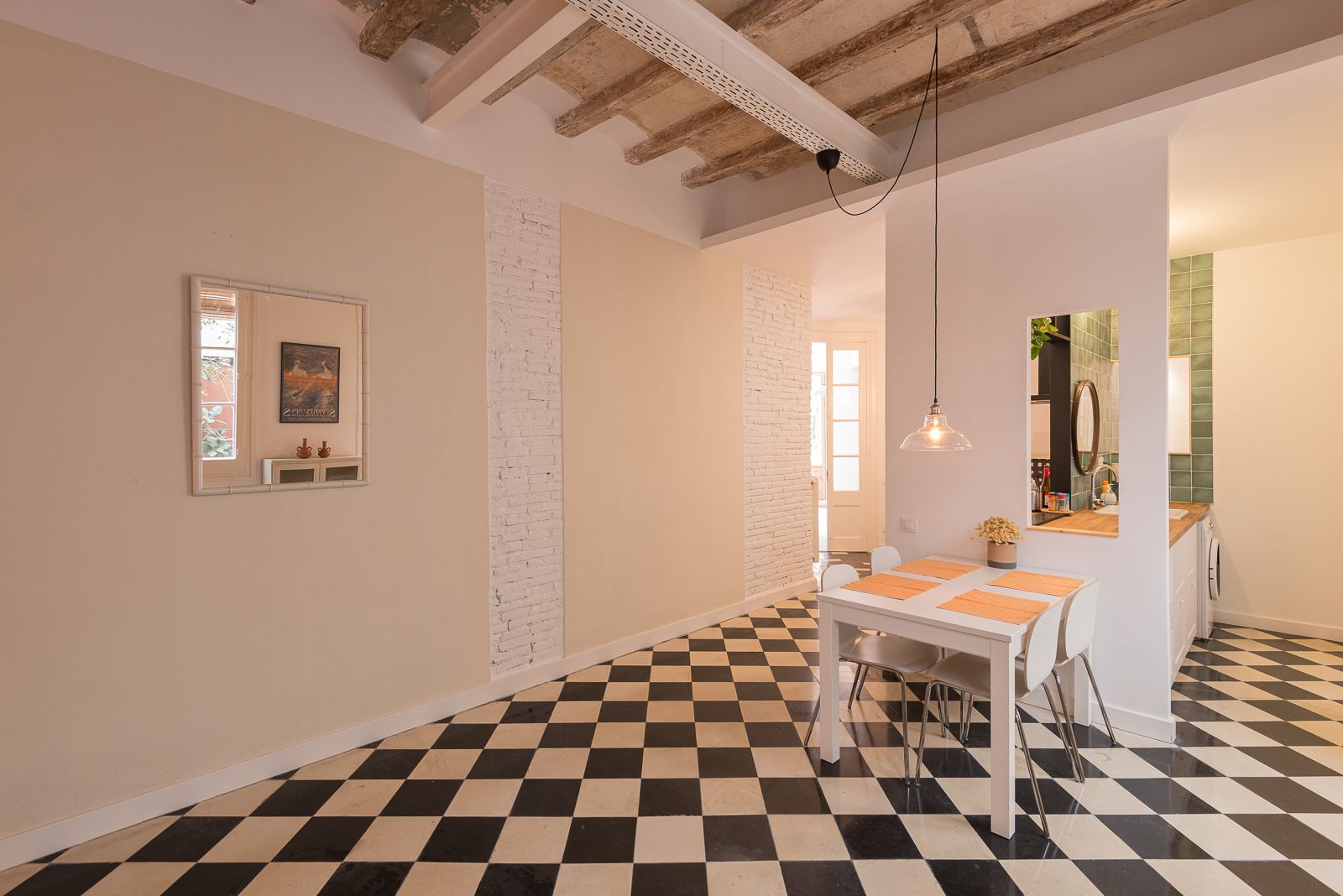 Zona de menjador moderna amb terra de quadres, bigues vistes i accents de maó blanc, amb una petita taula de menjador i llum penjant. Un mirall quadrat a la paret reflecteix una porta que s'obre a un pati
