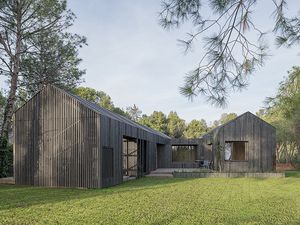 Exterior view of a modern house with two bodies, a pitched roof, dark wood cladding, large windows, and a spacious lawn surrounded by pine trees.