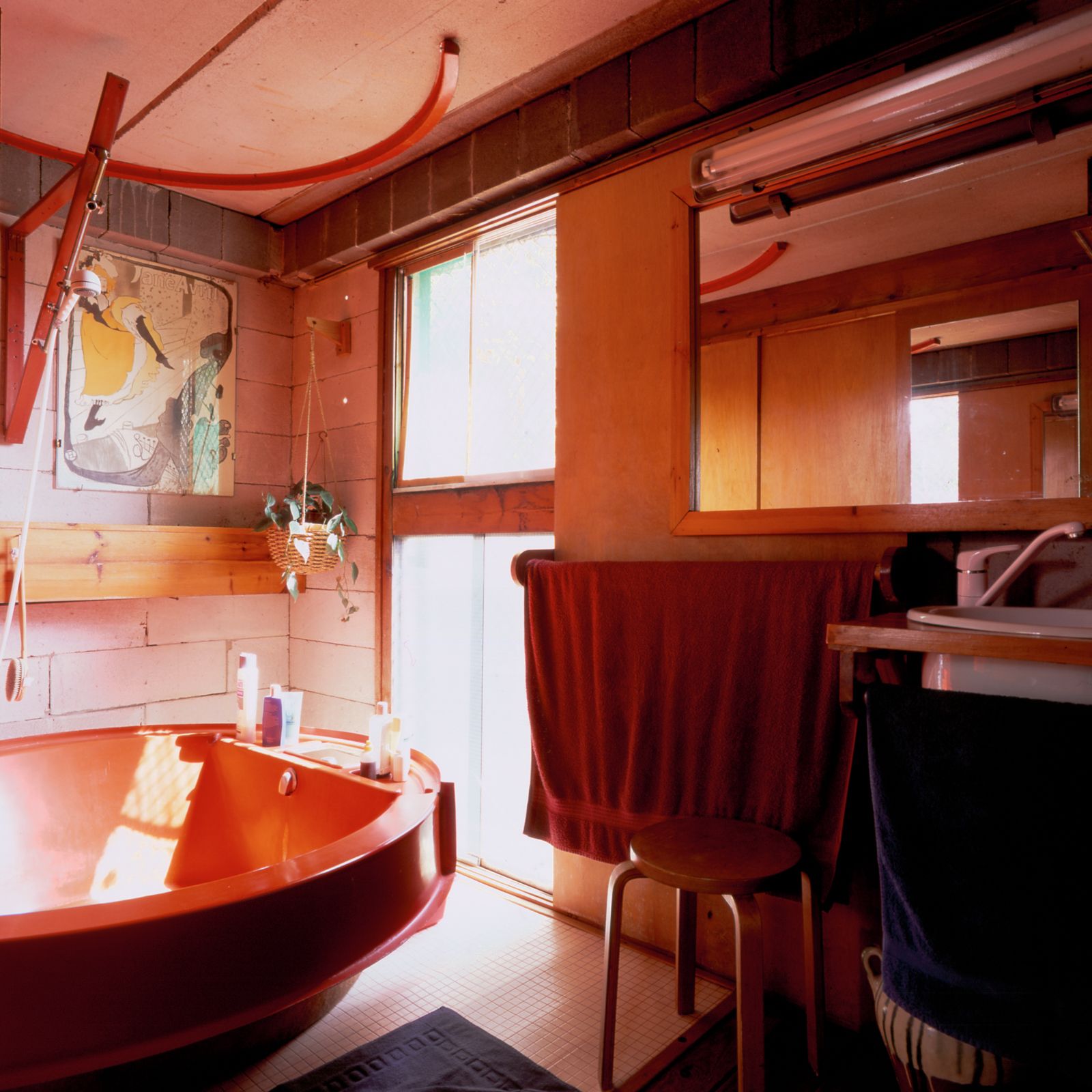 Bathroom with a red bathtub, wooden elements, a floor-to-ceiling window, and a variety of towels hanging below a mirror and a bathroom sink.