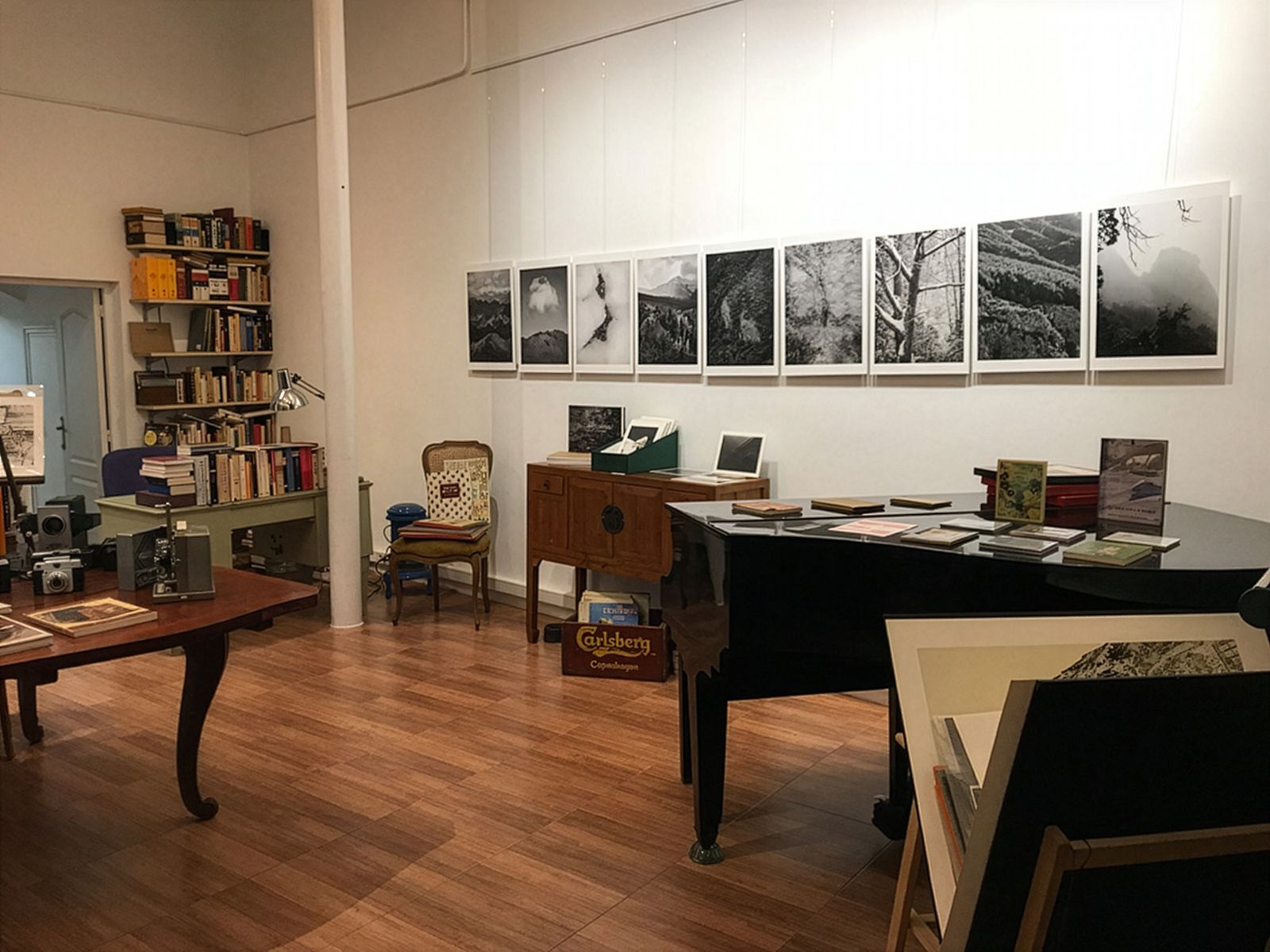 Un espai d'exposició amb fotografies en blanc i negre de paisatges muntanyosos exposades en una paret, un piano amb llibres i fullets a sobre, un escriptori amb càmeres antigues i prestatgeries al fons.