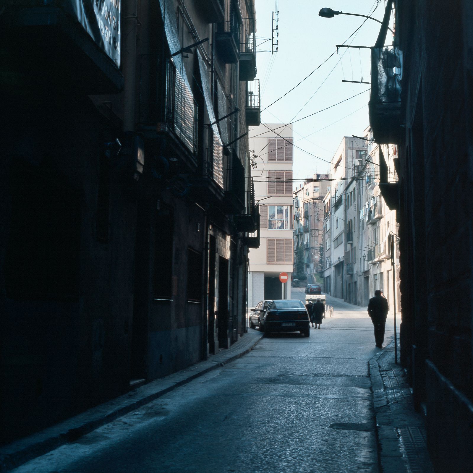 A narrow alleyway with shadowed buildings on both sides, leading to a brighter plaza and a new residential building in the background with a few pedestrians and parked cars.