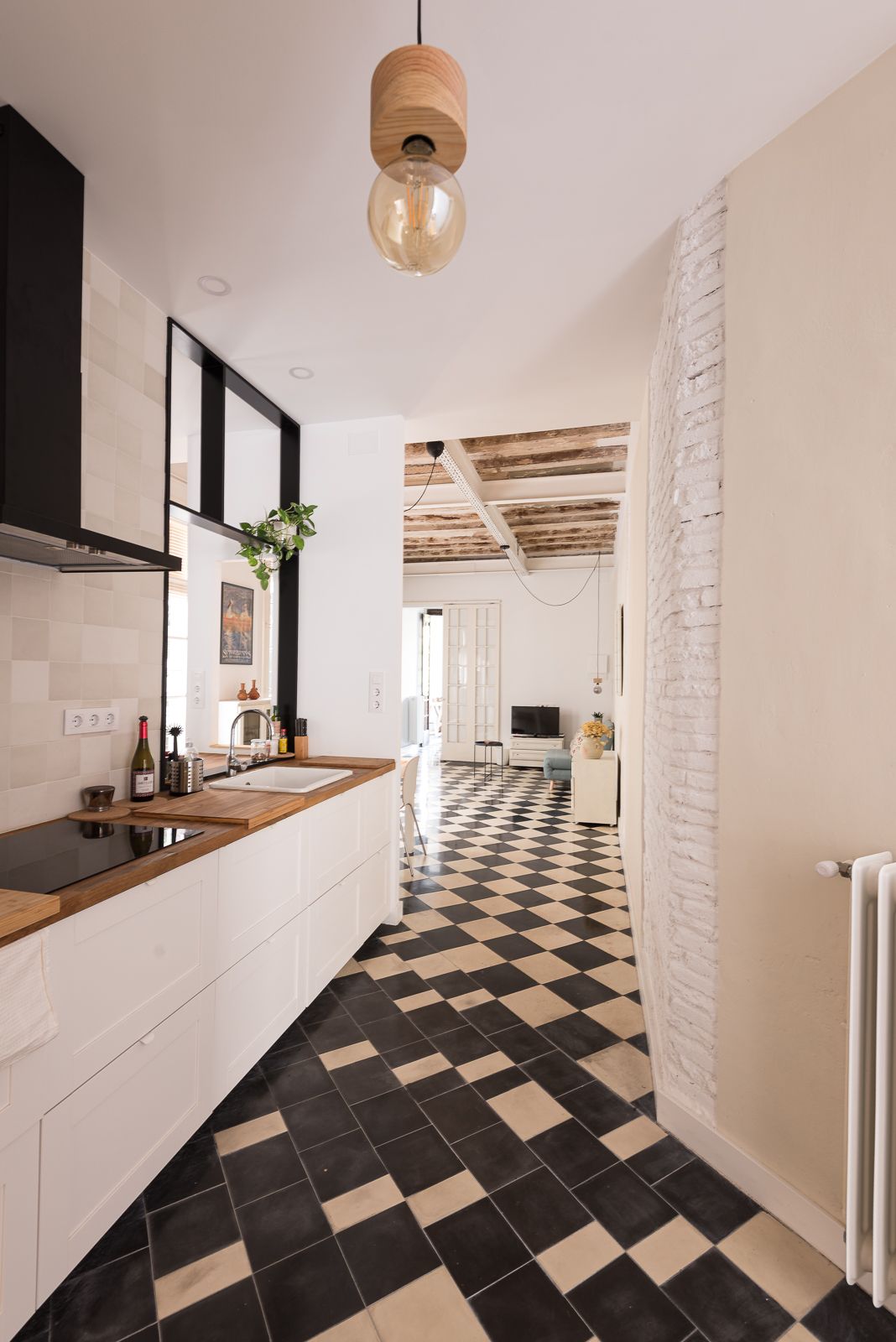 Cuina moderna amb terra de quadres, armaris blancs, taulells de fusta i un llum penjant elegant. Al fons, una gran zona d'estar amb terra de quadres i bigues de fusta.