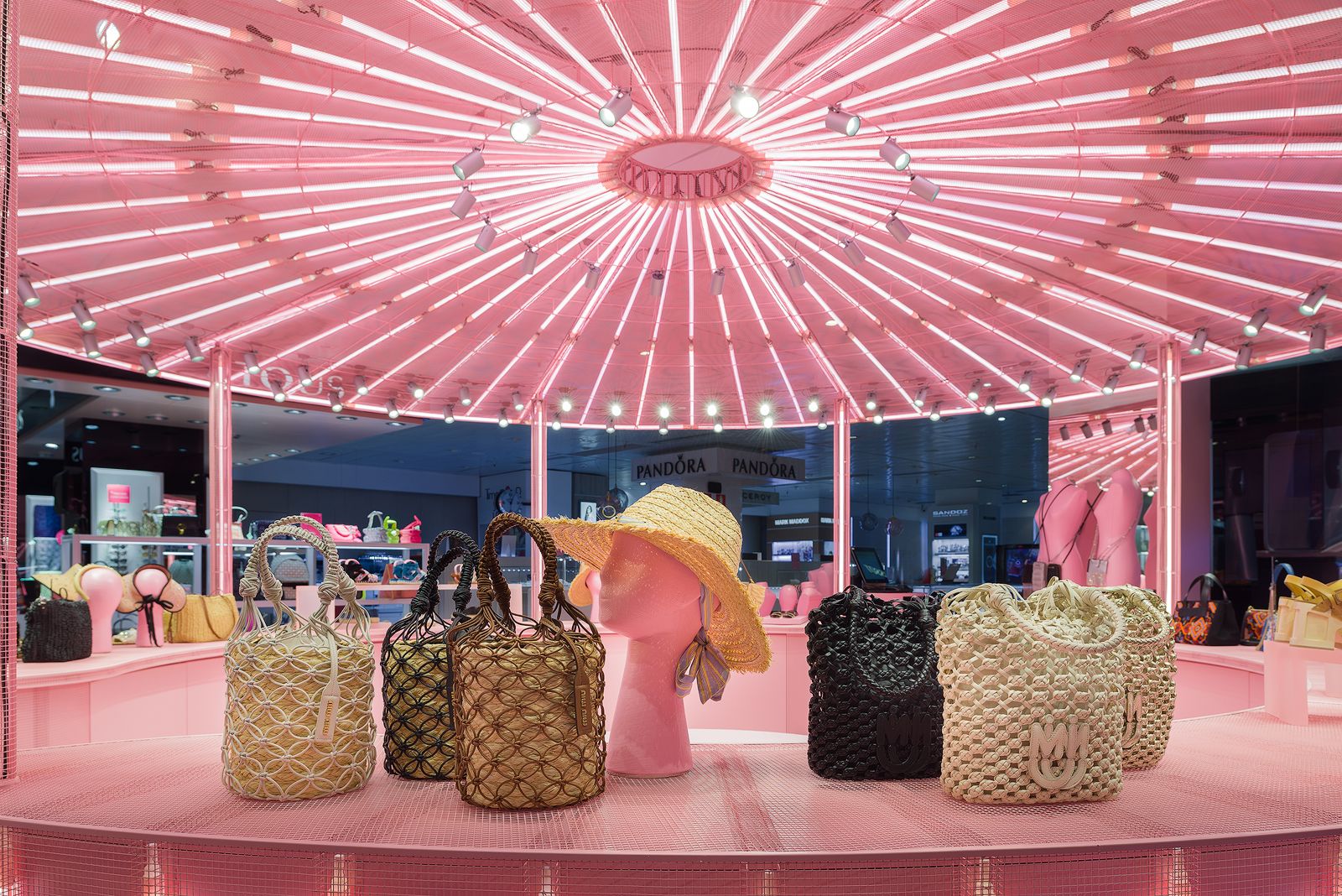 Woven bags and hats displayed under pink illuminated ceiling in Miu Miu's pop-up store.
