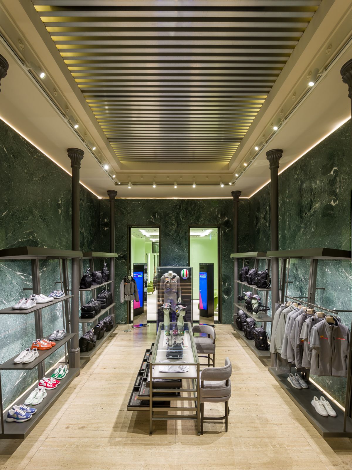 Luxurious boutique interior with green marble walls, elegant lighting, and black metal shelving displaying sneakers, bags, and gray apparel. A central glass table showcases accessories, while a doorway at the back leads to another section.
