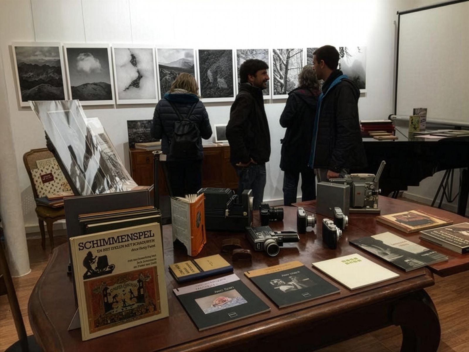 Un grup de persones veient fotografies en blanc i negre exposades en un espai d'exposició acollidor. La sala disposa d'una taula de fusta amb càmeres antigues, llibres i altres objectes. Les fotografies mostren paisatges muntanyosos i altres escenes naturals.