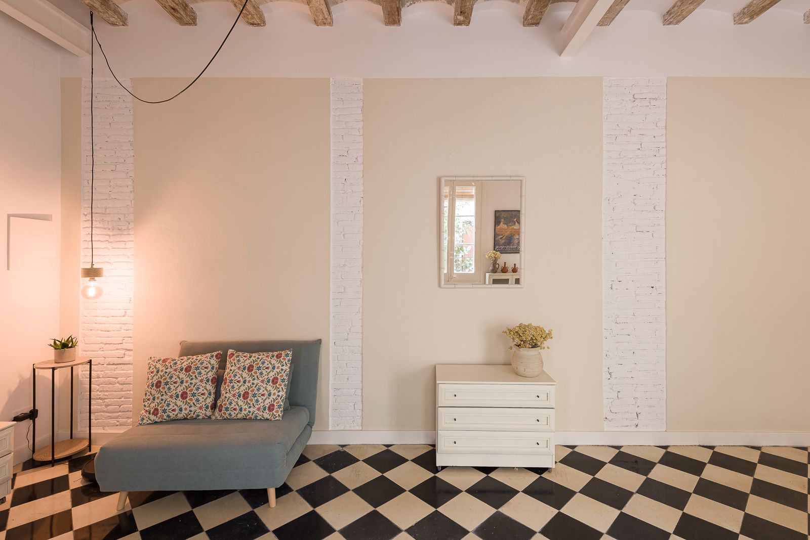 Racó acollidor amb una butaca blava, coixins florals, una tauleta, una calaixera blanca, un mirall quadrat a la paret que reflecteix la porta al pati i rajoles de terra de quadres.