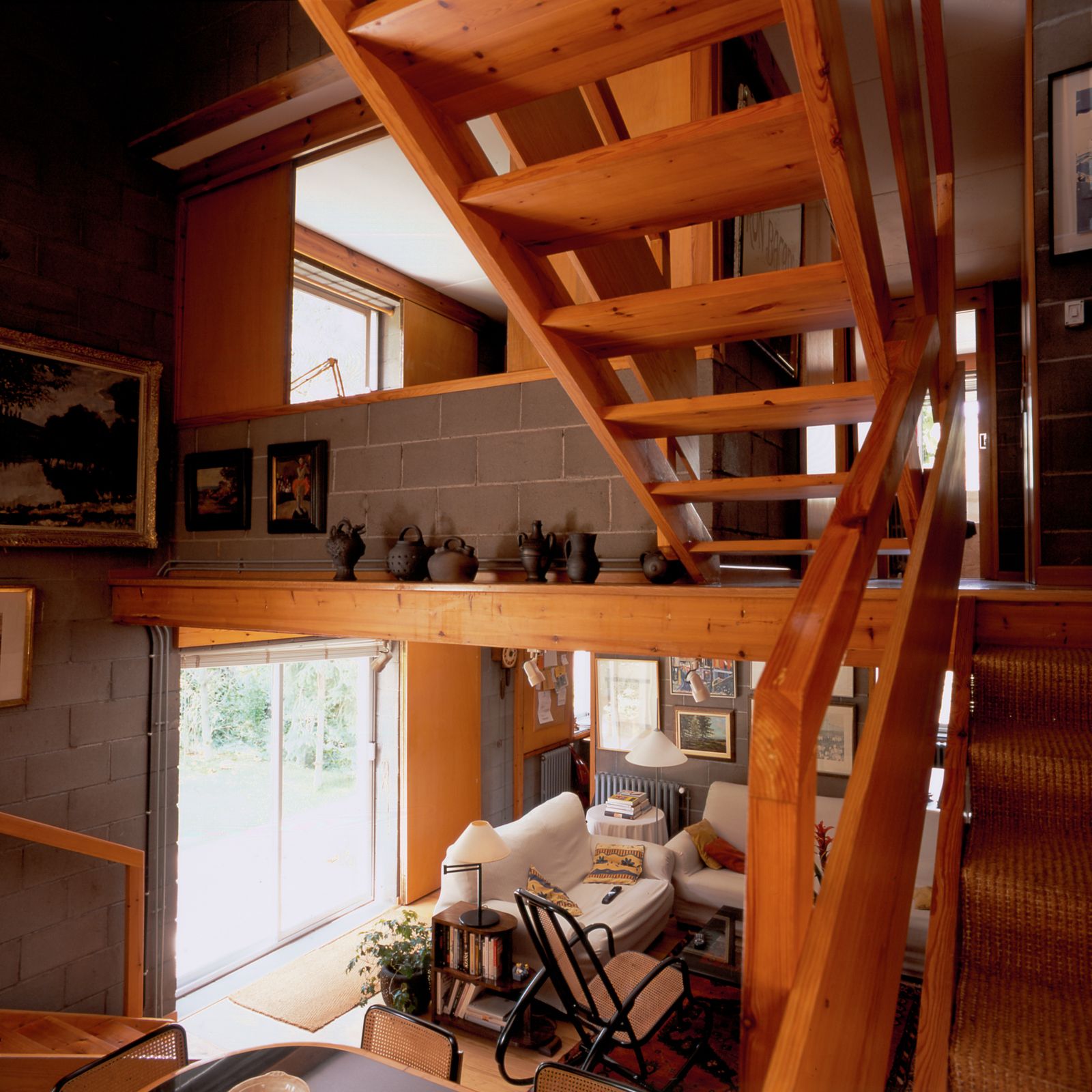 Vista interior que mostra una escala de fusta, una sala d'estar acollidora amb sofàs blancs, una cadira gronxadora, una catifa i objectes decoratius en prestatges i parets. L'habitació té parets de blocs de formigó i una gran porta corredera que deixa entrar llum natural.