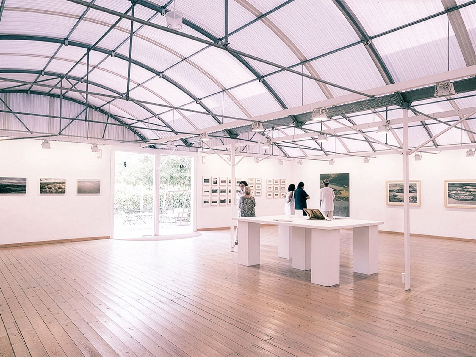 Personas viendo obras de arte enmarcadas en una sala de exposición espaciosa y luminosa con suelos de madera y un techo alto y curvo.