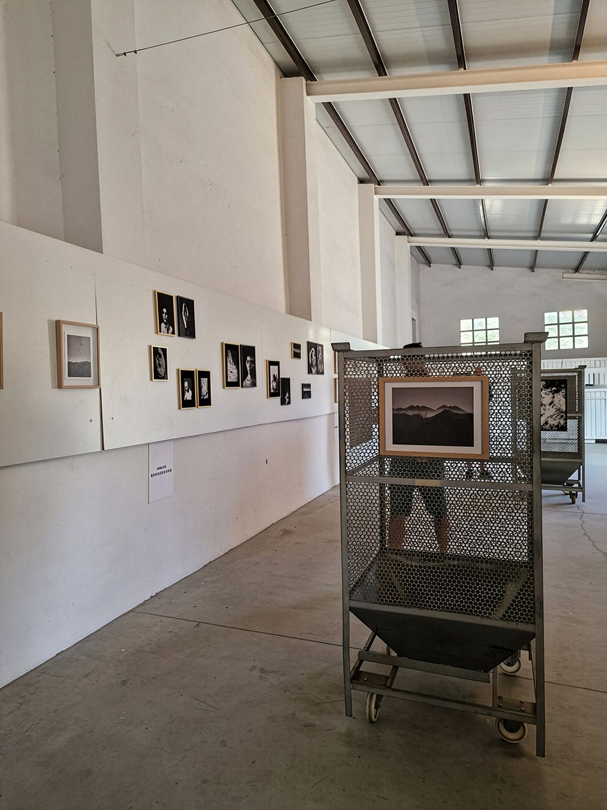 Interior de una galería de arte con techos altos y vigas expuestas. Las paredes blancas muestran una serie de fotografías enmarcadas. En primer plano, un soporte de exhibición de malla metálica sobre ruedas sostiene una fotografía de paisaje enmarcada. El suelo de hormigón y las ventanas de estilo industrial sugieren un espacio de almacén convertido.