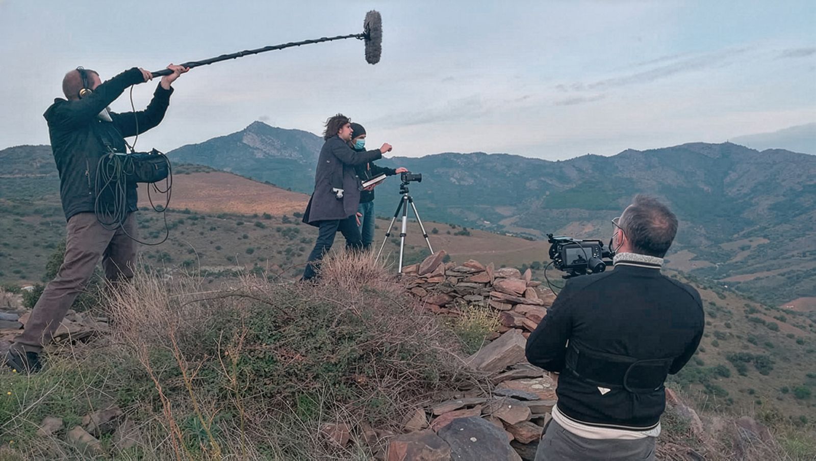 Un equipo de filmación rodando en una ladera con un telón de fondo montañoso panorámico. El equipo incluye un camarógrafo y una persona sosteniendo un micrófono boom, están filmando a dos personas montando un trípode con una cámara.