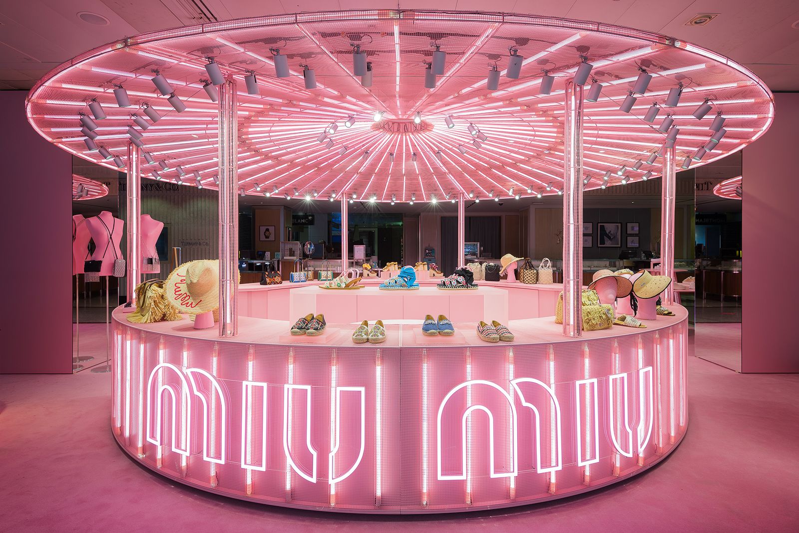Illuminated pink Miu Miu pop-up store display with handbags, shoes, and hats under a canopy of bright pink lights.