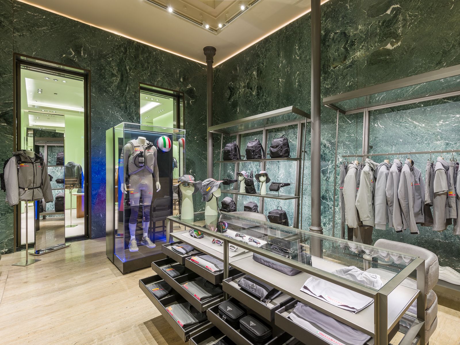 Luxury Prada boutique with green marble walls, showcasing designer sneakers, black bags, and accessories on sleek metal shelving. A glass table displays eyewear and hats, while a mannequin in a performance outfit is encased in a glass display.