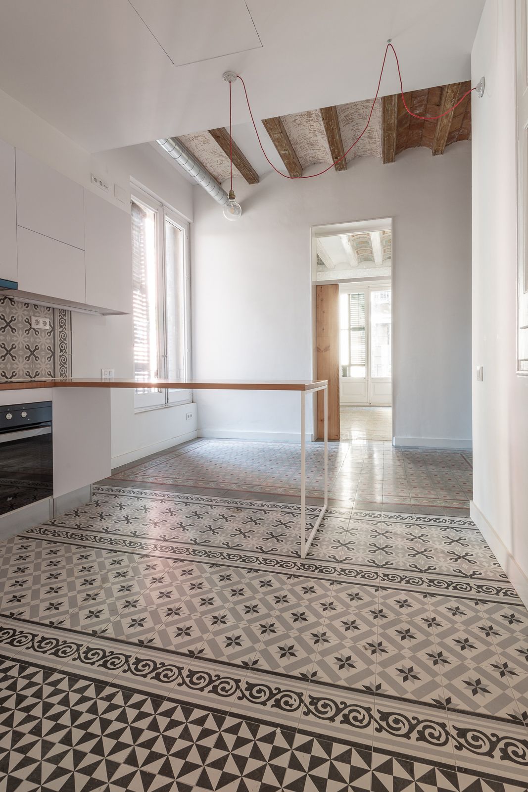 Bright kitchen with patterned floor tiles, exposed wooden beams, and a minimalist work table.