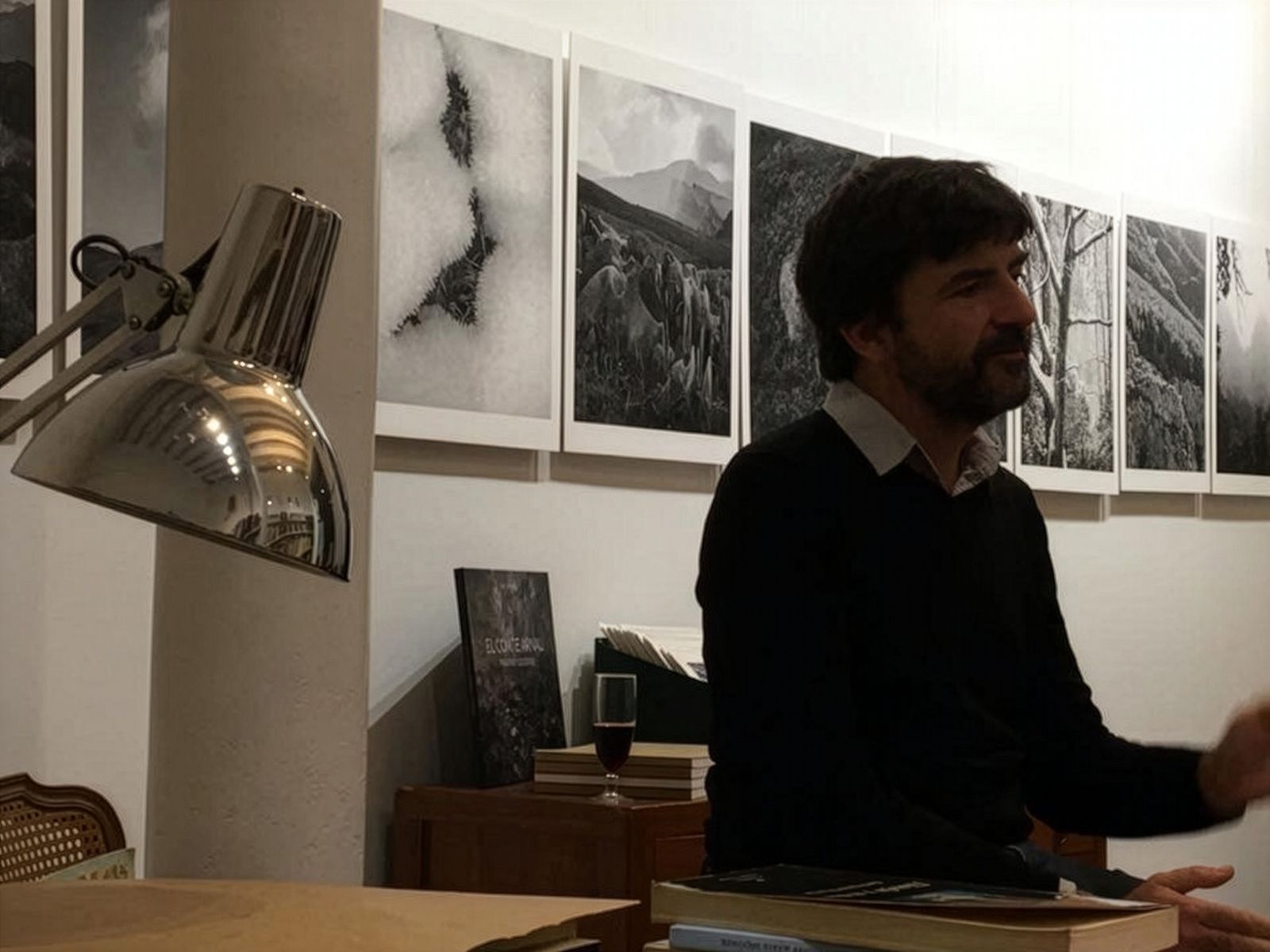 Un home seu davant d'una mostra de fotografies en blanc i negre de paisatges muntanyosos en un espai d'exposició acollidor. Hi ha un llum d'escriptori a prop i una copa de vi sobre un moble al darrere.
