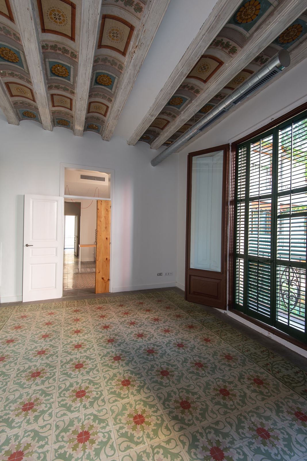 Room with ornate ceiling beams and patterned floor tiles, featuring a partially open wooden door and shuttered window.