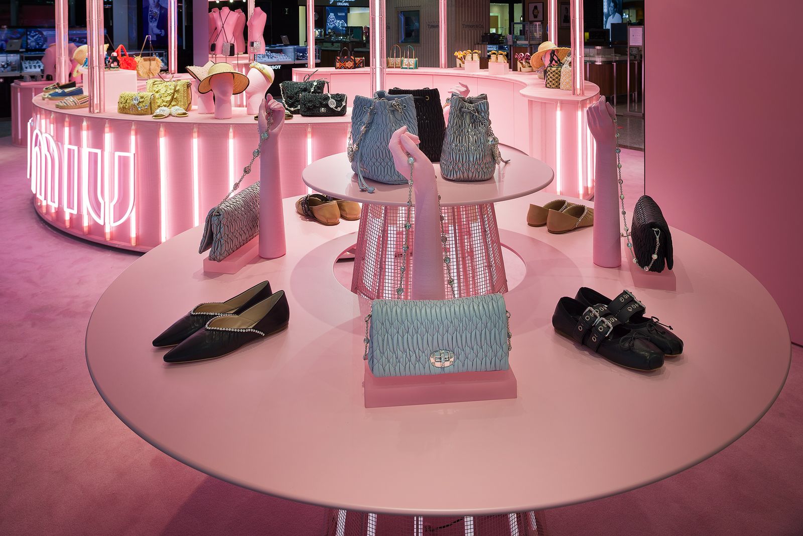 Close-up of a pink-themed Miu Miu pop-up store display, featuring handbags and shoes arranged on a two-tier round table with a conic base illuminated from within by neon lighting. Standing on the table, three pink mannequin hands showcasing accessories.