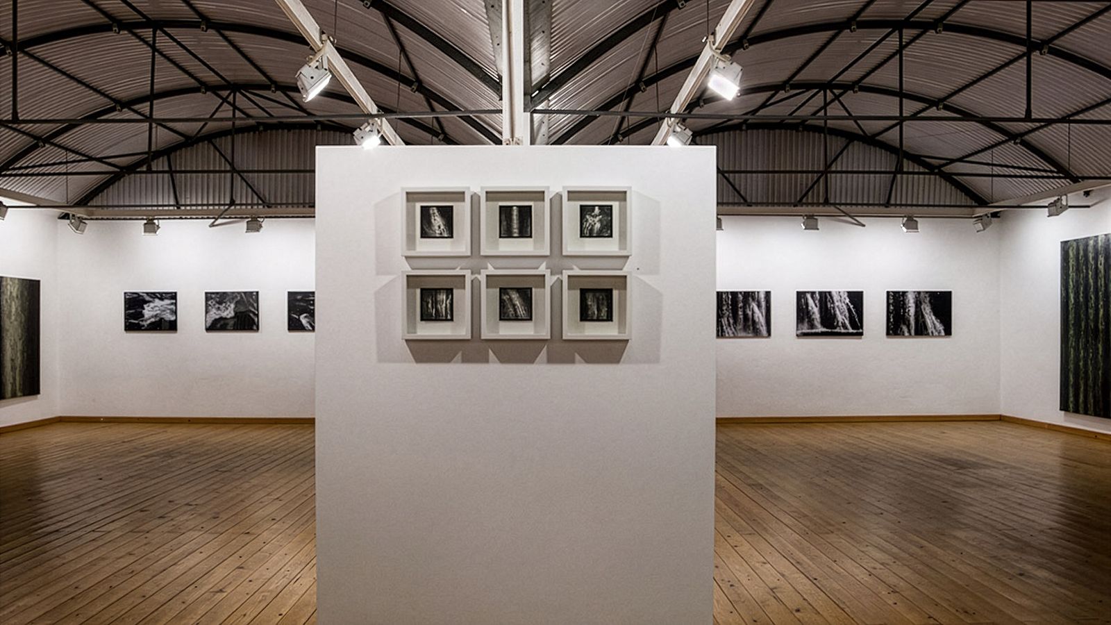 Interior de galería con obras de arte enmarcadas en las paredes, exposición central de marcos más pequeños, suelos de madera y techo metálico abovedado.