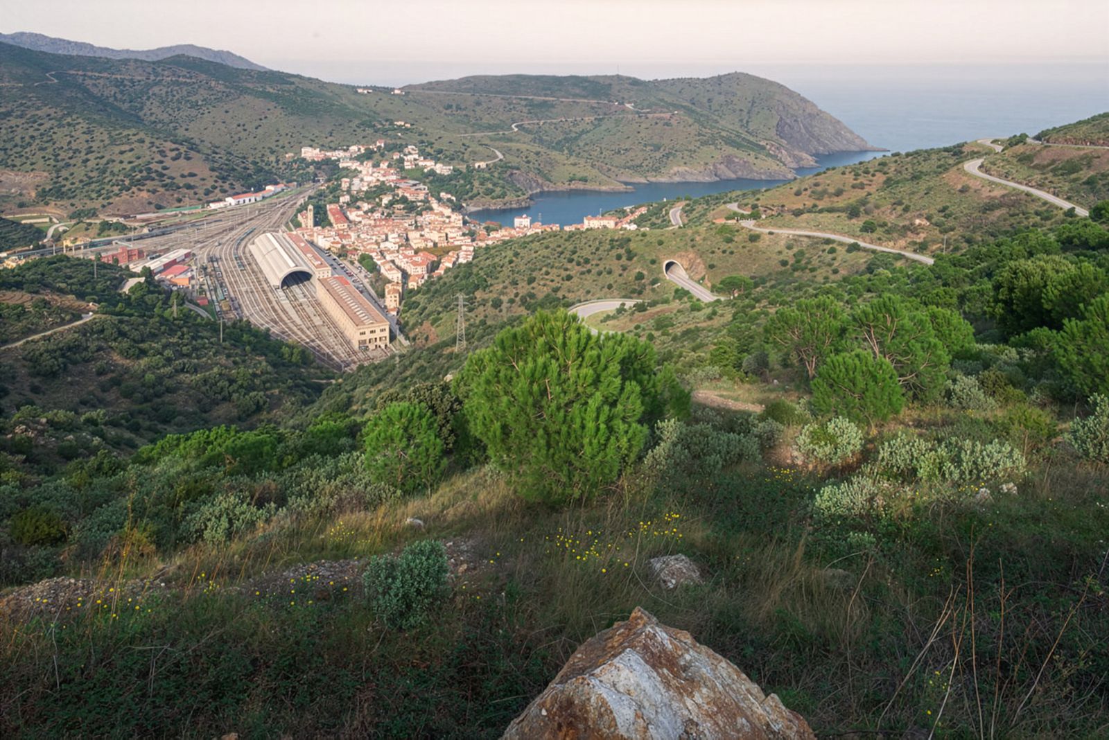 Una vista panorámica de un pueblo costero con una enorme estación de tren con múltiples vías terminales, rodeado de colinas verdes y con vistas a una bahía serena.