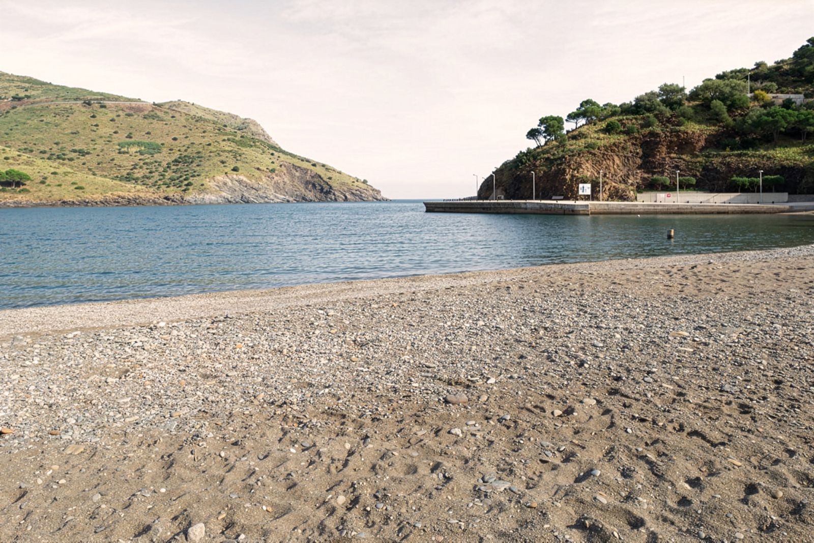 Una playa tranquila de guijarros con aguas azules tranquilas, rodeada de colinas verdes y afloramientos rocosos.