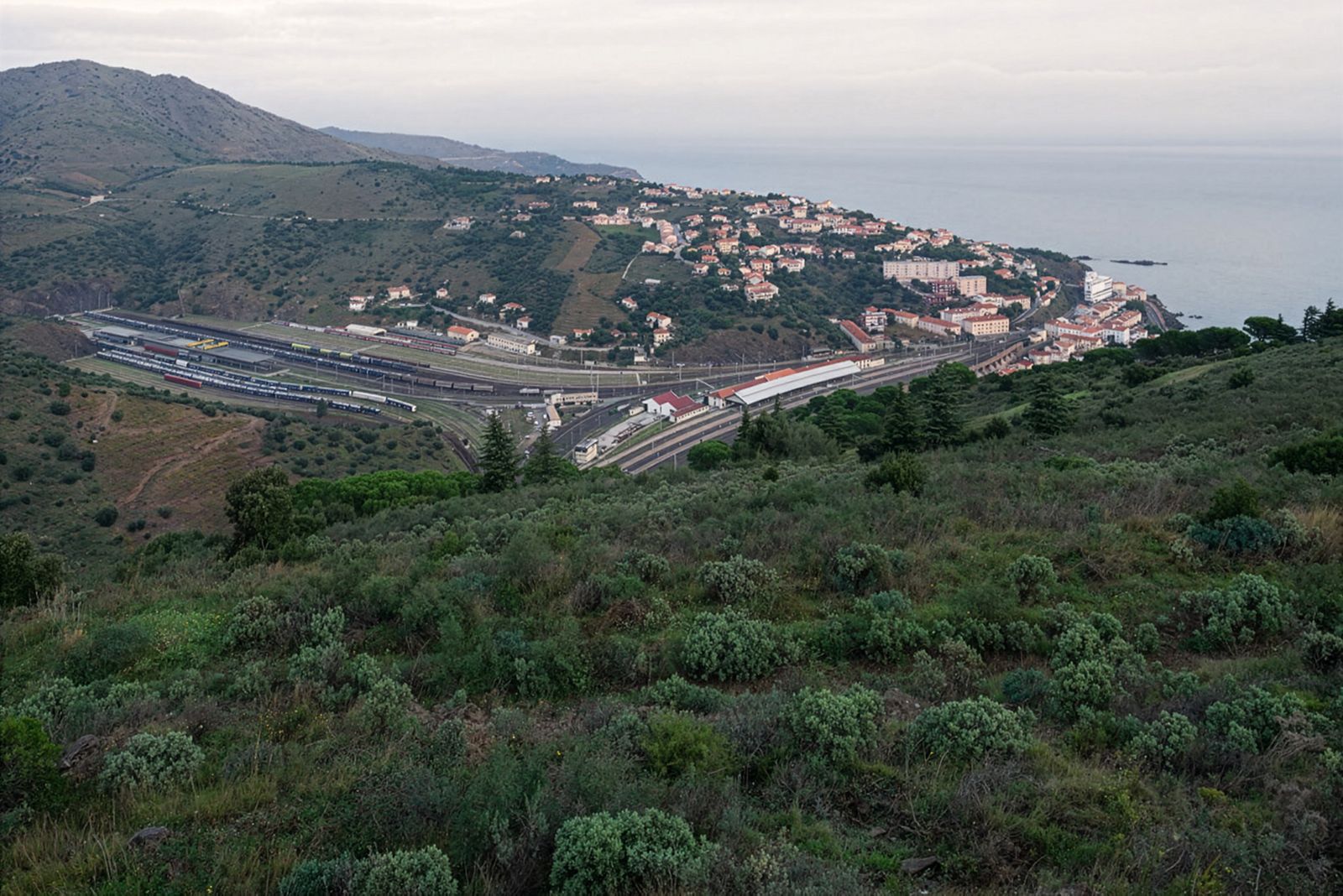 Una vista de un pueblo costero con una gran estación de tren con múltiples vías terminales que atraviesan un valle y casas dispersas en la ladera de la colina, extendiéndose hacia el mar.