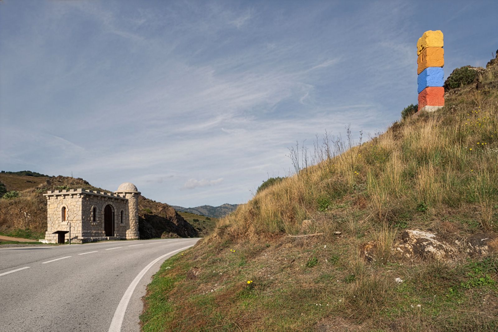Una carretera sinuosa con un minúsculo edificio de piedra que parece un castillo a la izquierda y un pilar apilado colorido a la derecha, con un telón de fondo de colinas ondulantes bajo un cielo azul.